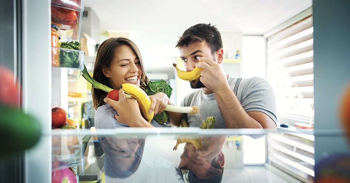 Two happy young adults holding produce in front of a refrigerator.