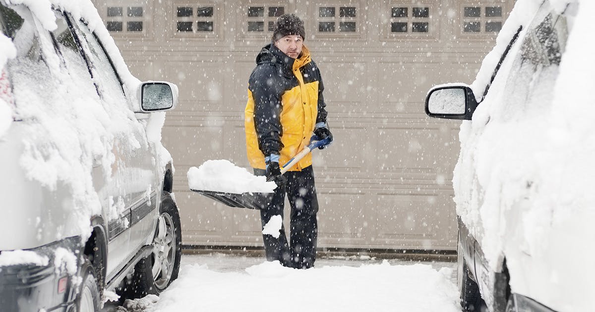 Snow falls heavily. A man in a yellow and black coat stands between two parked cars holding a big shovel full of snow. 