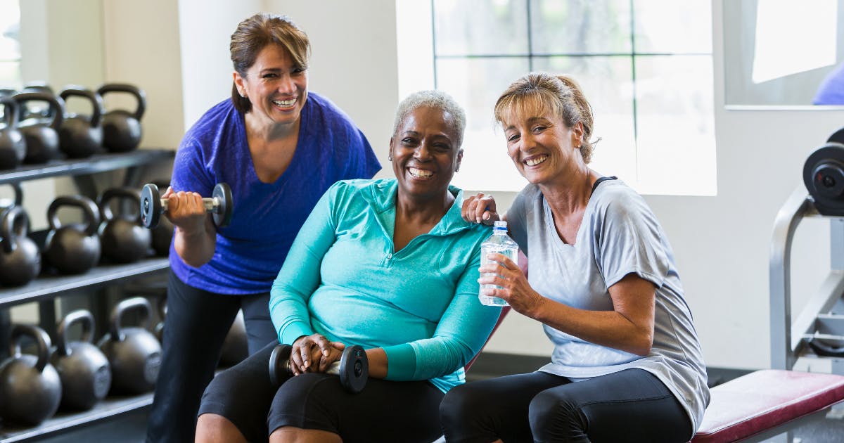 Three smiling women on a weight bench at a gym.