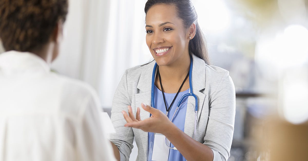 A doctor in a white coat with a blue stethoscope smiles and chats with a patient. 