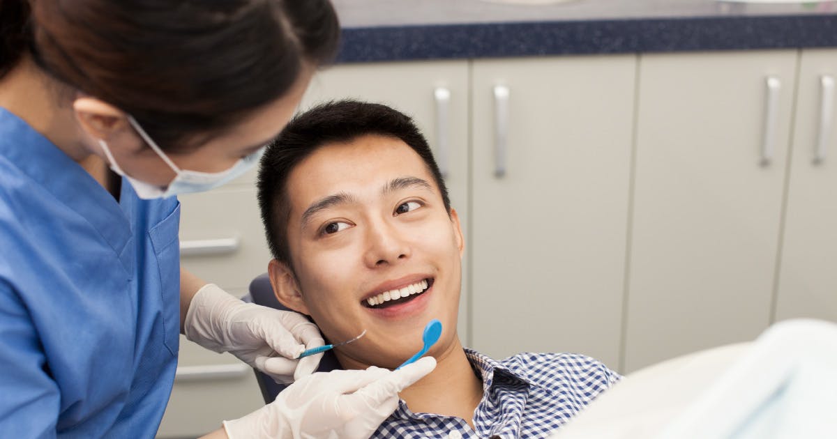 A dental hygienist holds a pick and mirror near a smiling young man in a dental chair