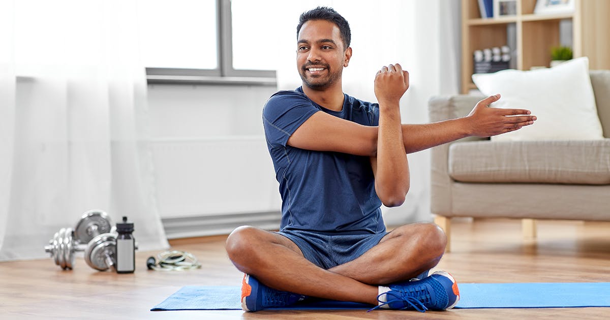 A man sits on an exercise mat and stretches his arm.