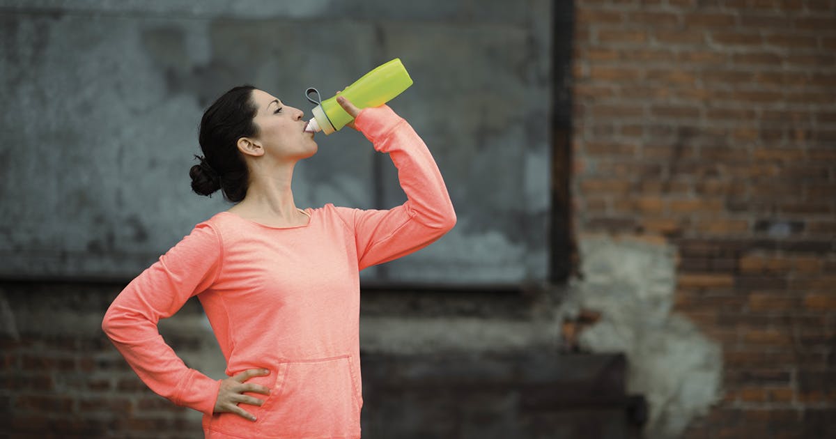 A woman standing outside in athletic gear takes a break to drink from her water bottle.