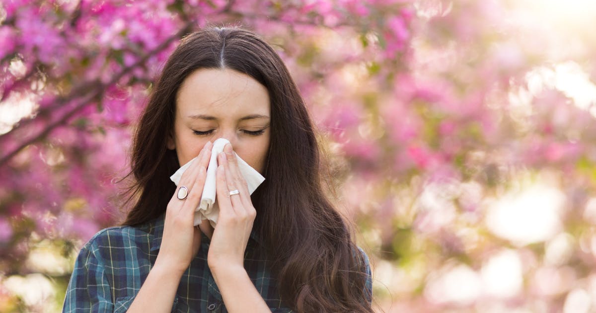 A woman stands in front of a cherry tree while holding a tissue to her nose.