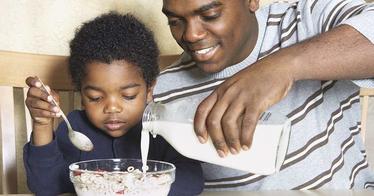  A smiling Black father adds milk to his son's cereal.