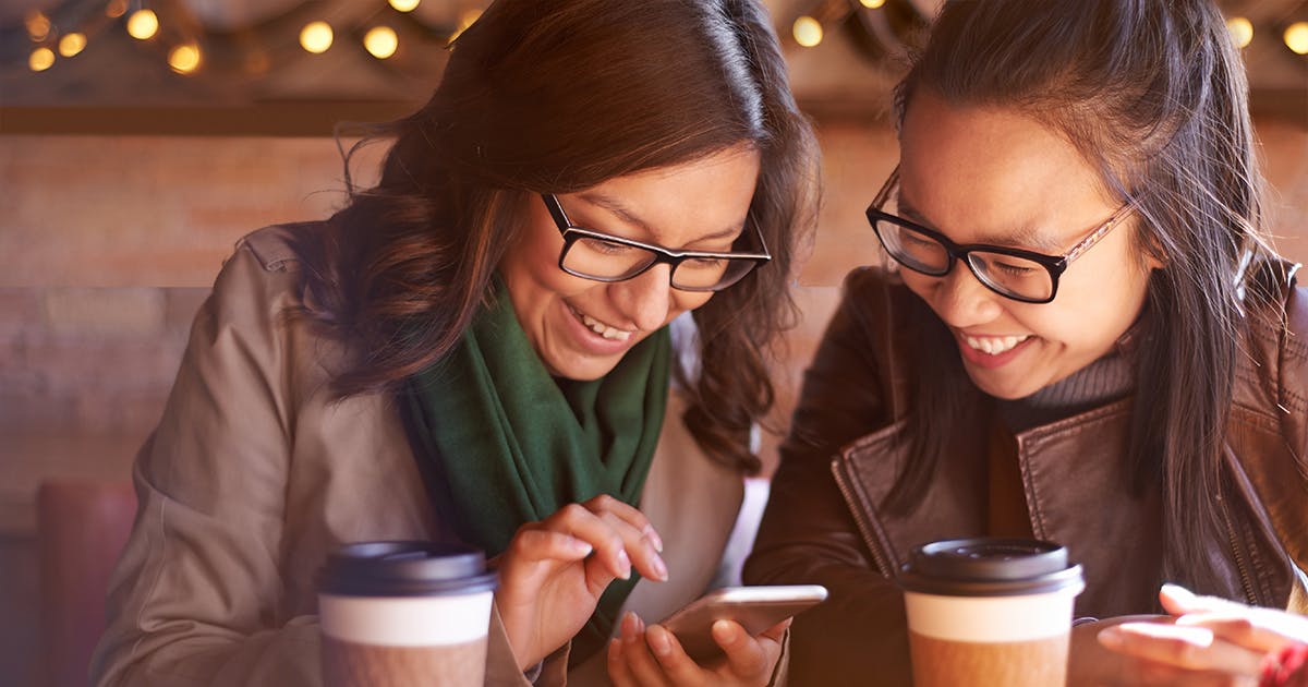 A pair of women in a coffee shop laugh while looking at a phone.
