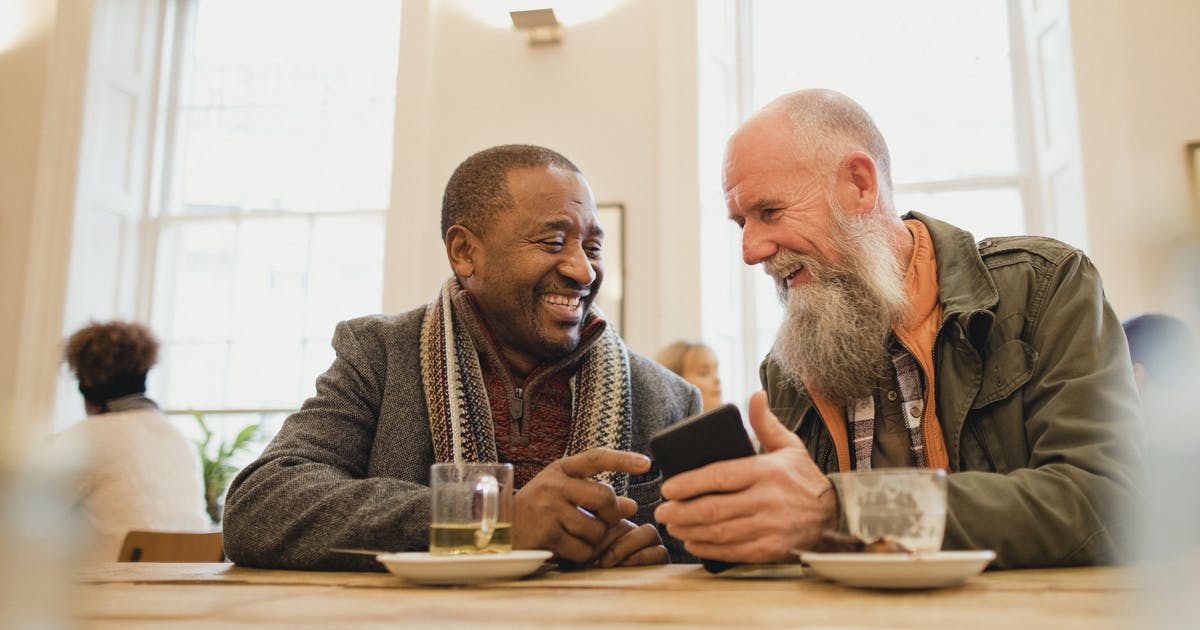 Two men in a coffee shop look at a smartphone and smile at each other.