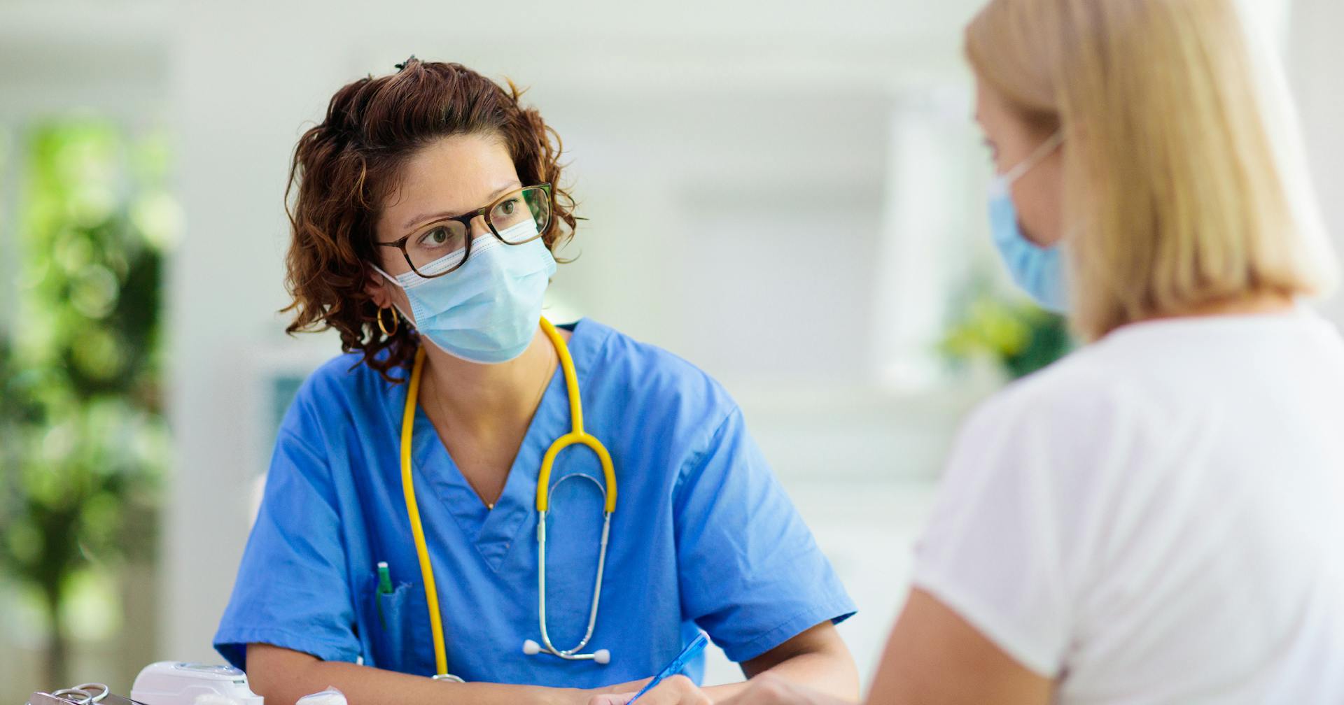 Health care provider takes notes while she talks with a patient (both wearing masks).