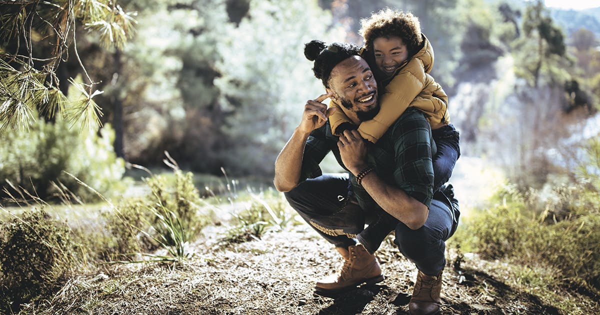 A man crouched on a hiking trail with a smiling child on his back.