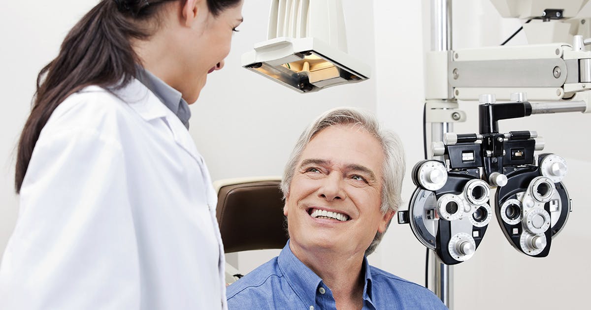 A man with silver hair sits in an eye exam chair, smiling up at his doctor, a woman in a white coat. 
