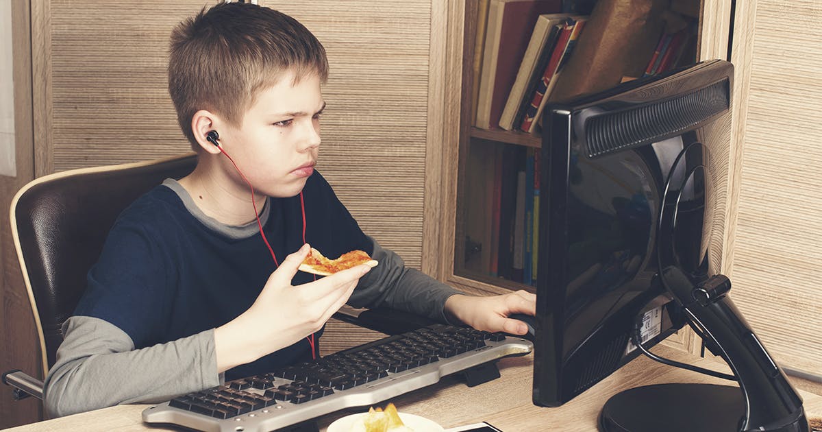 A boy sits in front of a computer eating pizza.