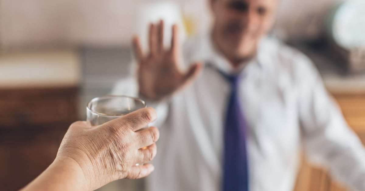 A hand holds out a glass to a man gesturing no in the background.