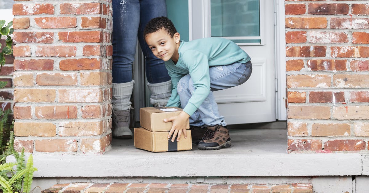 A boy picks up packages on his doorstep, with his mother’s legs visible behind him.