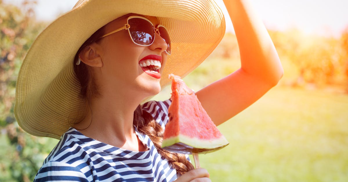 A smiling woman in a hat and sunglasses holding a slice of watermelon on a stick.