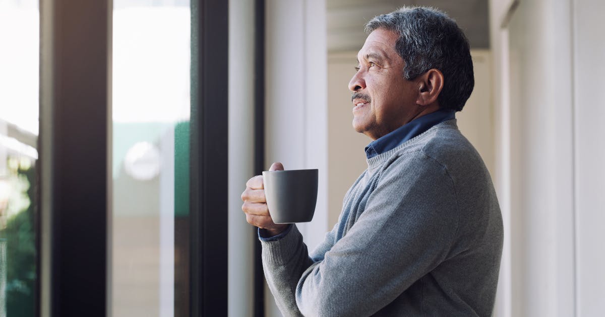 A man holds a mug and looks out a window.
