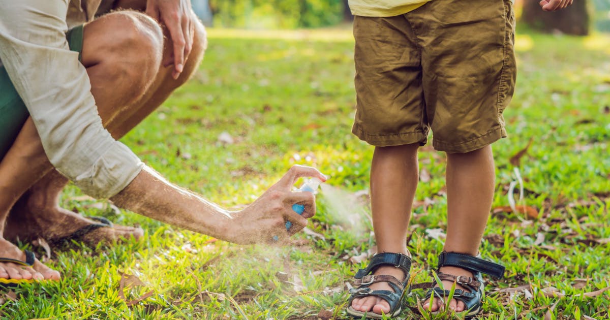 An adult squats to spray mosquito repellent on a child’s ankles.