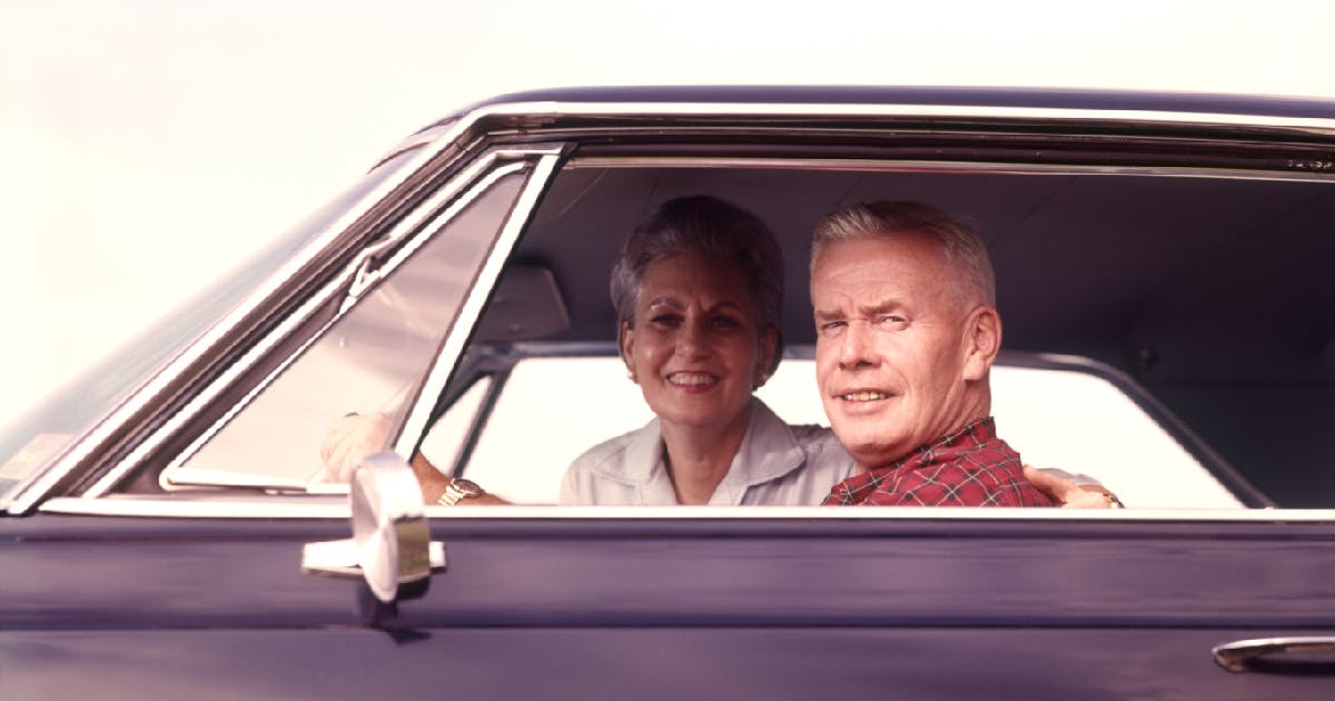 A smiling older couple seen through the window of an older model car.