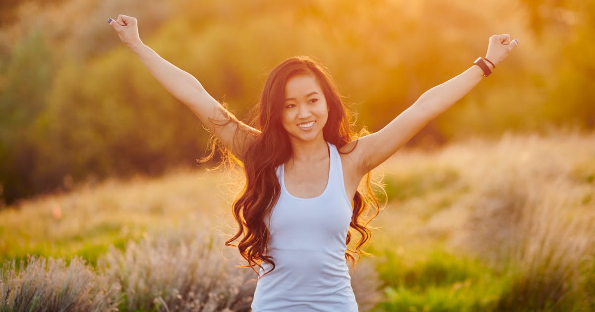 A smiling young woman in a tank top stands in a sunlit field, arms raised in a wide pose